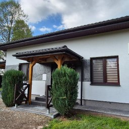 REMONTY & WNĘTRZA Piotr Piestrzyński - Biała elewacja domu z brązowymi oknami i drewnianym zadaszeniem nad wejściem. Widoczny klimatyzator i zadbana zieleń przed budynkiem.