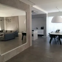 Nowoczesne wnętrze z dużym lustrem na ścianie z efektem betonu architektonicznego, odbijającym salon z szarą sofą i minimalistycznym oświetleniem.