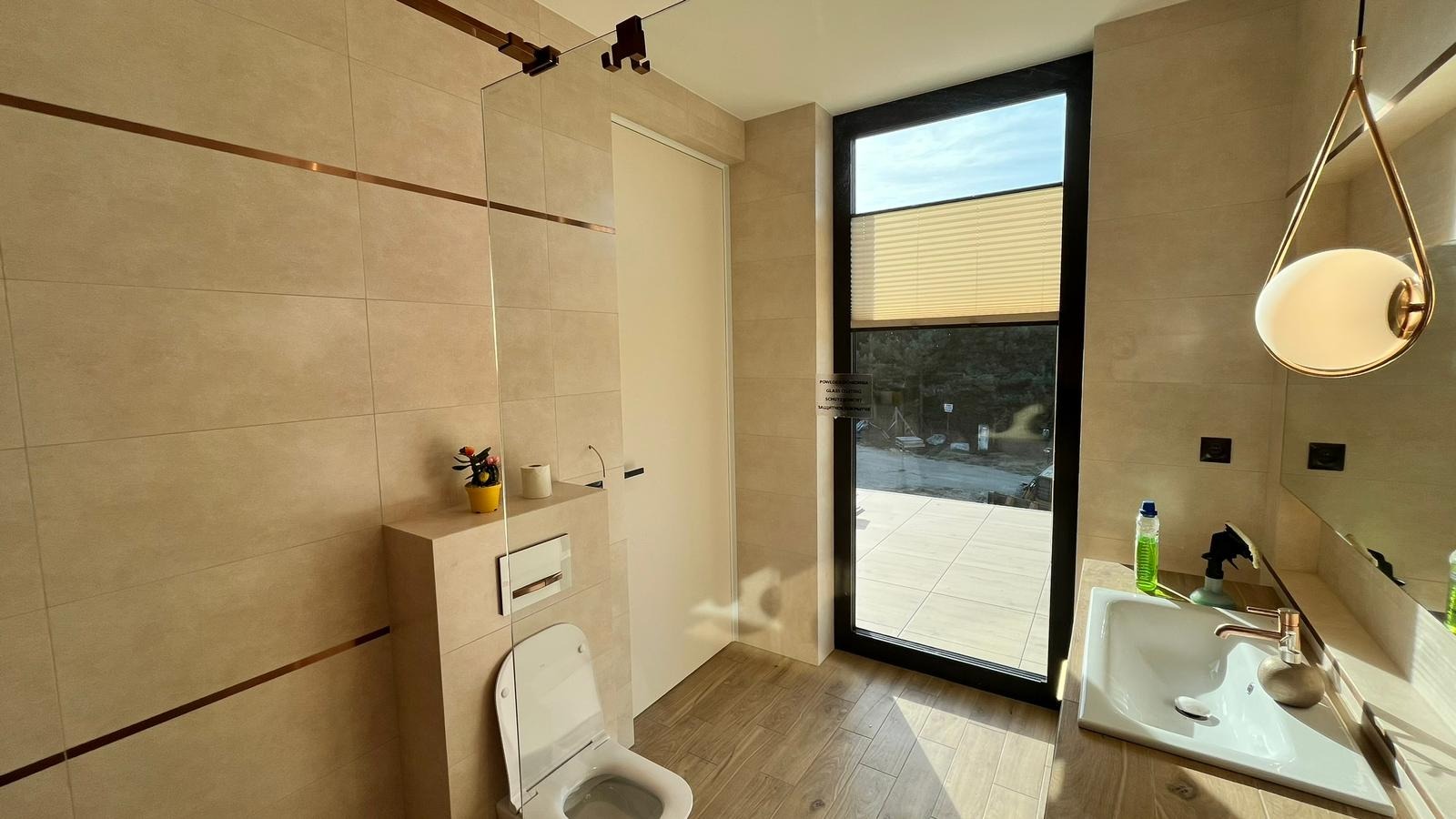 Nowoczesna łazienka z beżowymi płytkami, drewnopodobną podłogą, kabiną prysznicową ze szklaną ścianką i wiszącą lampą w kształcie kuli.