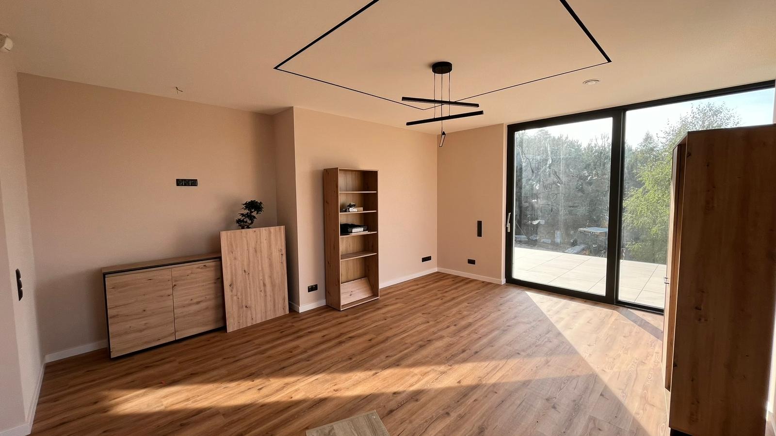 Jasne wnętrze pokoju z panelami podłogowymi, beżowymi ścianami, nowoczesnym oświetleniem sufitowym i przeszkleniami z widokiem na taras.