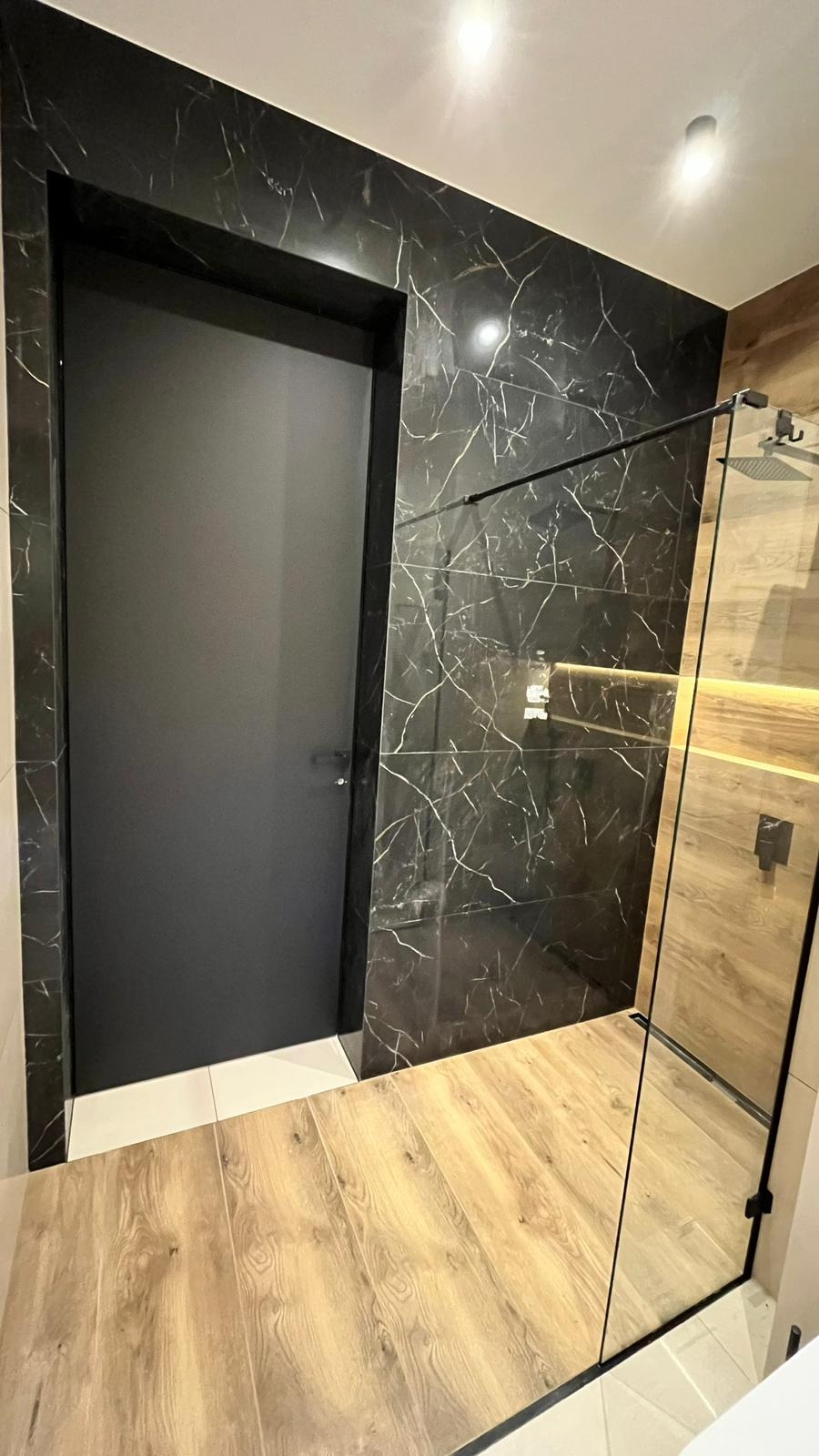 Nowoczesna łazienka z czarnymi płytkami imitującymi marmur, drewnopodobną podłogą i kabiną prysznicową ze szklaną ścianką oraz czarnymi detalami, obok czarne drzwi.