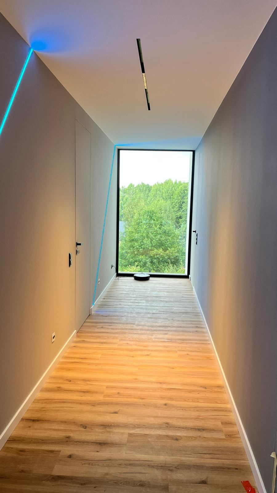 Wnętrze nowoczesnego korytarza z drewnopodobną podłogą, dużym oknem wychodzącym na zieleń, minimalistycznym oświetleniem sufitowym i dekoracyjnym niebieskim paskiem LED w rogu ściany.