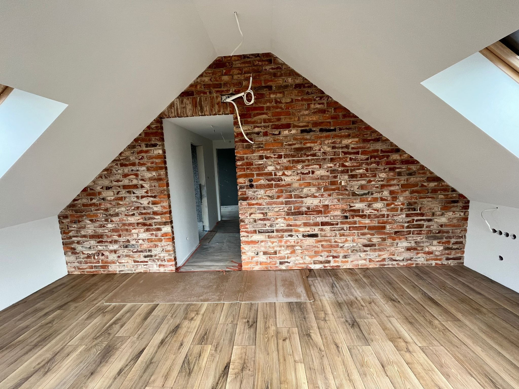 Poddasze z wyeksponowaną ścianą z czerwonej cegły, drewniana podłoga w trakcie układania, widoczne przewody elektryczne i otwór drzwiowy do kolejnego pomieszczenia.