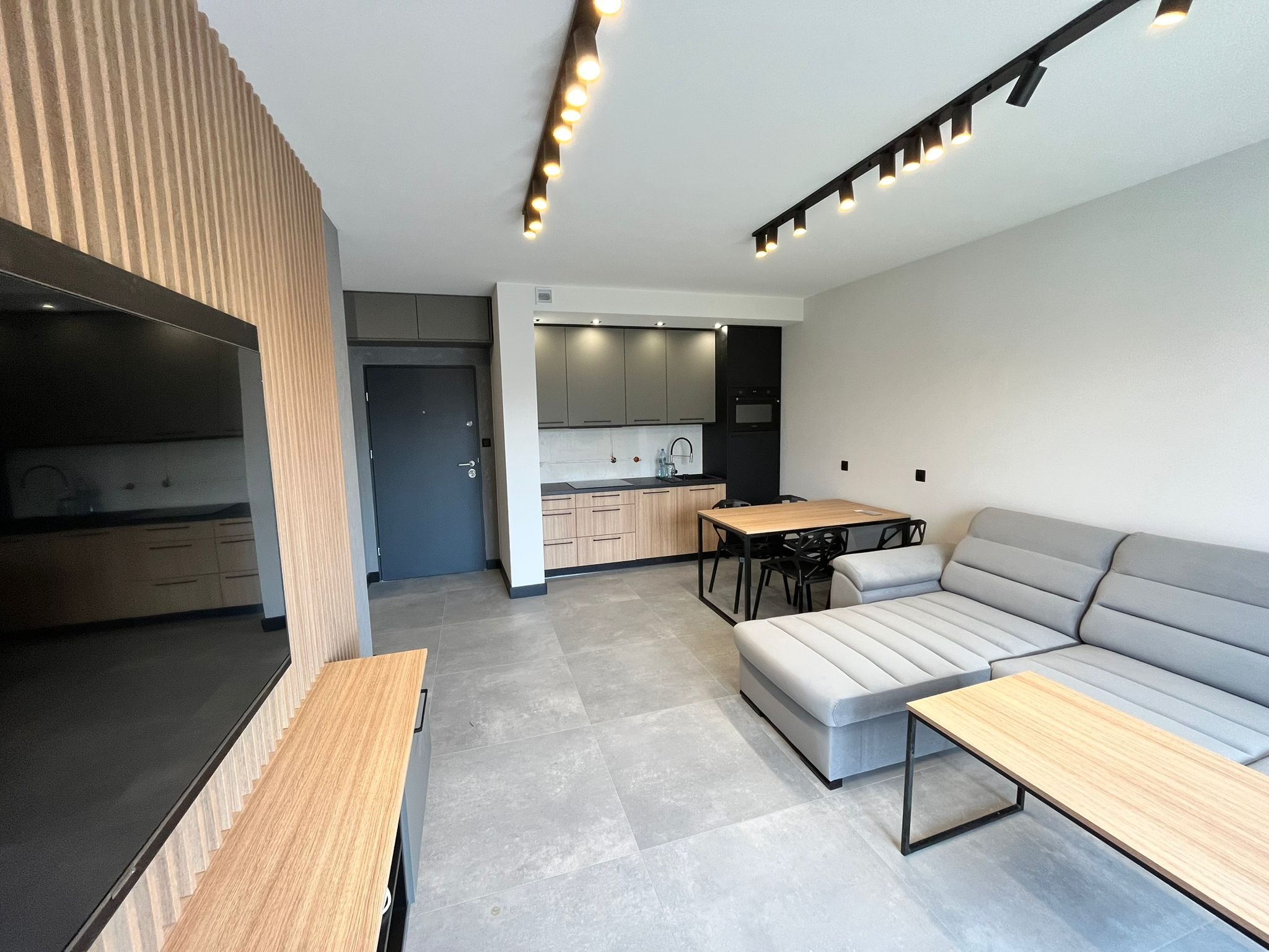 Nowoczesne wnętrze mieszkania z szarą sofą narożną, drewnianym stolikiem kawowym, ścianą z drewnianych lameli, aneksem kuchennym w odcieniach szarości i beżu oraz oświetleniem szynowym.