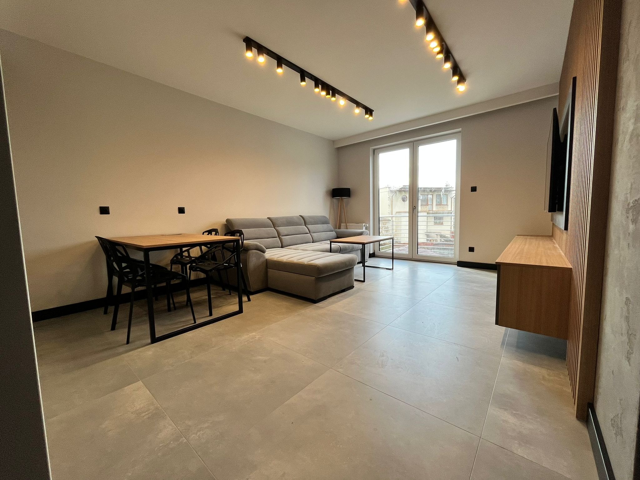 Nowoczesny salon z szarą sofą narożną, drewnianym stołem i czarnymi krzesłami, oświetleniem punktowym na suficie i dużymi oknami balkonowymi.