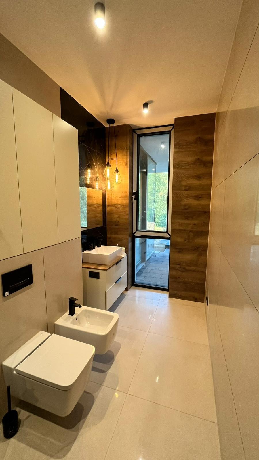 Nowoczesna łazienka z białą ceramiką, drewnopodobnymi płytkami na ścianie i wiszącymi lampami z żarówkami Edisona, widok na zewnątrz przez duże okno.