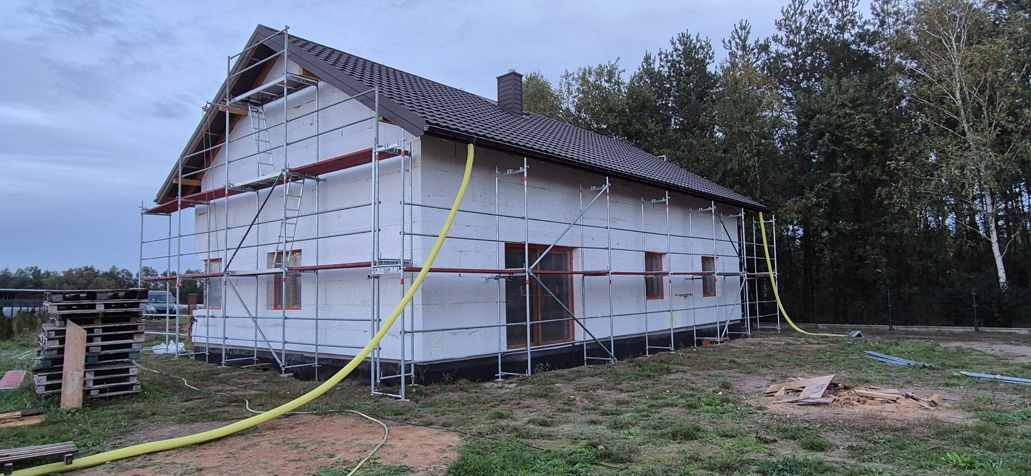 Dom w trakcie ocieplania styropianem, z rusztowaniem. Żółty wąż na elewacji. Widoczne palety i fragment lasu w tle.
