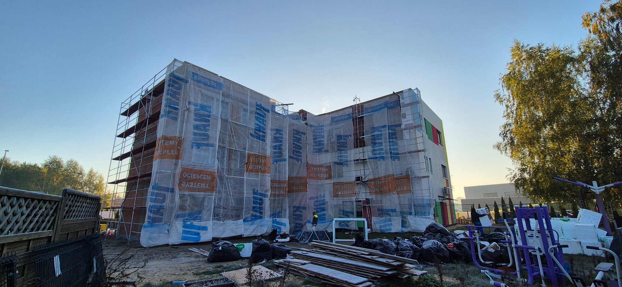 Budynek w trakcie ocieplania, osłonięty siatką z logo Knauf, rusztowania, widoczne materiały budowlane. Prace w toku, fragment elewacji z kolorem.