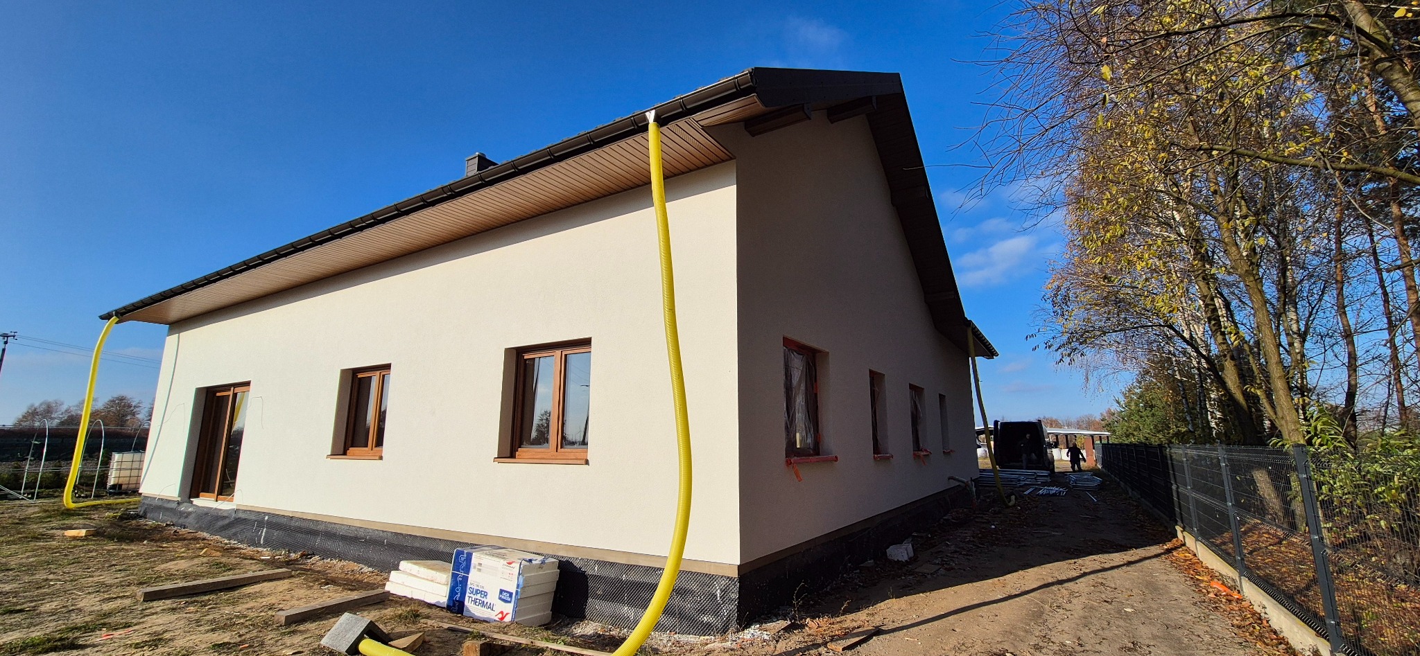 Jasny dom jednorodzinny po tynkowaniu, z widocznymi oknami zabezpieczonymi folią. Na pierwszym planie żółty wąż i styropian. Błękitne niebo w tle.