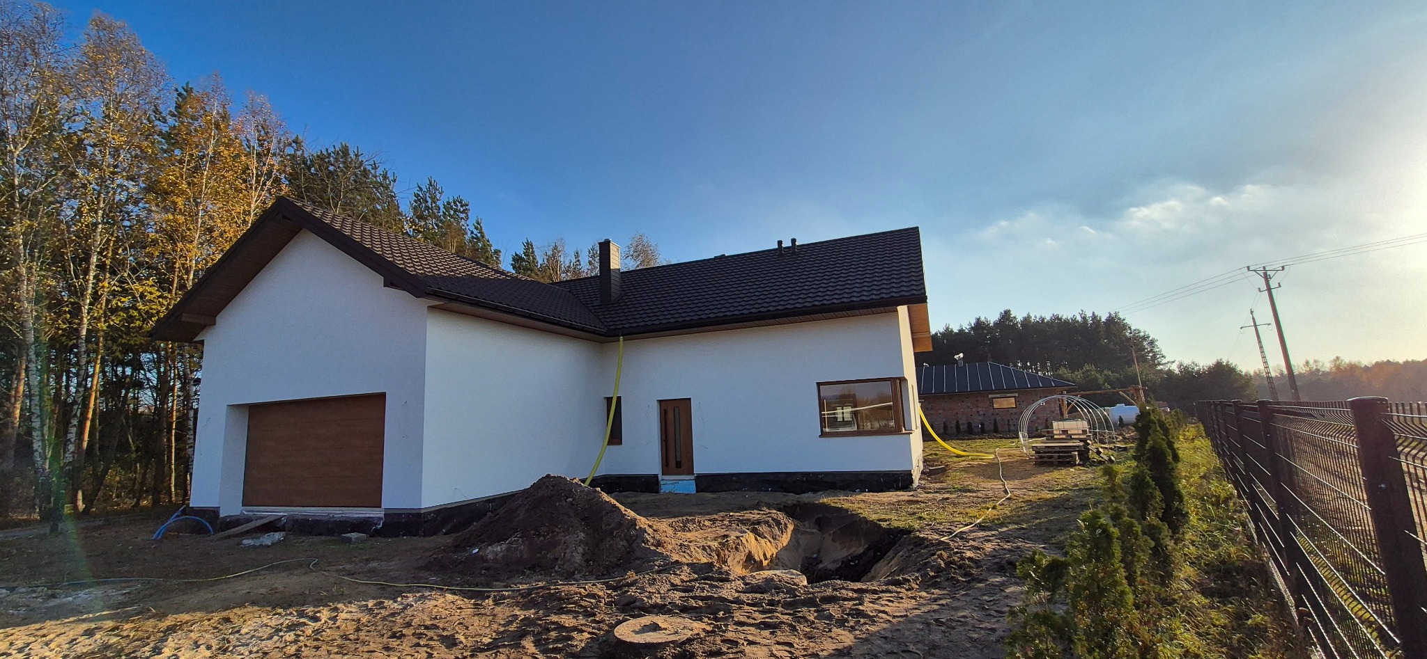Nowoczesny dom jednorodzinny z białą elewacją, ciemnym dachem i brązowymi oknami, w trakcie prac wykończeniowych, z wykopem i stertą ziemi na pierwszym planie.