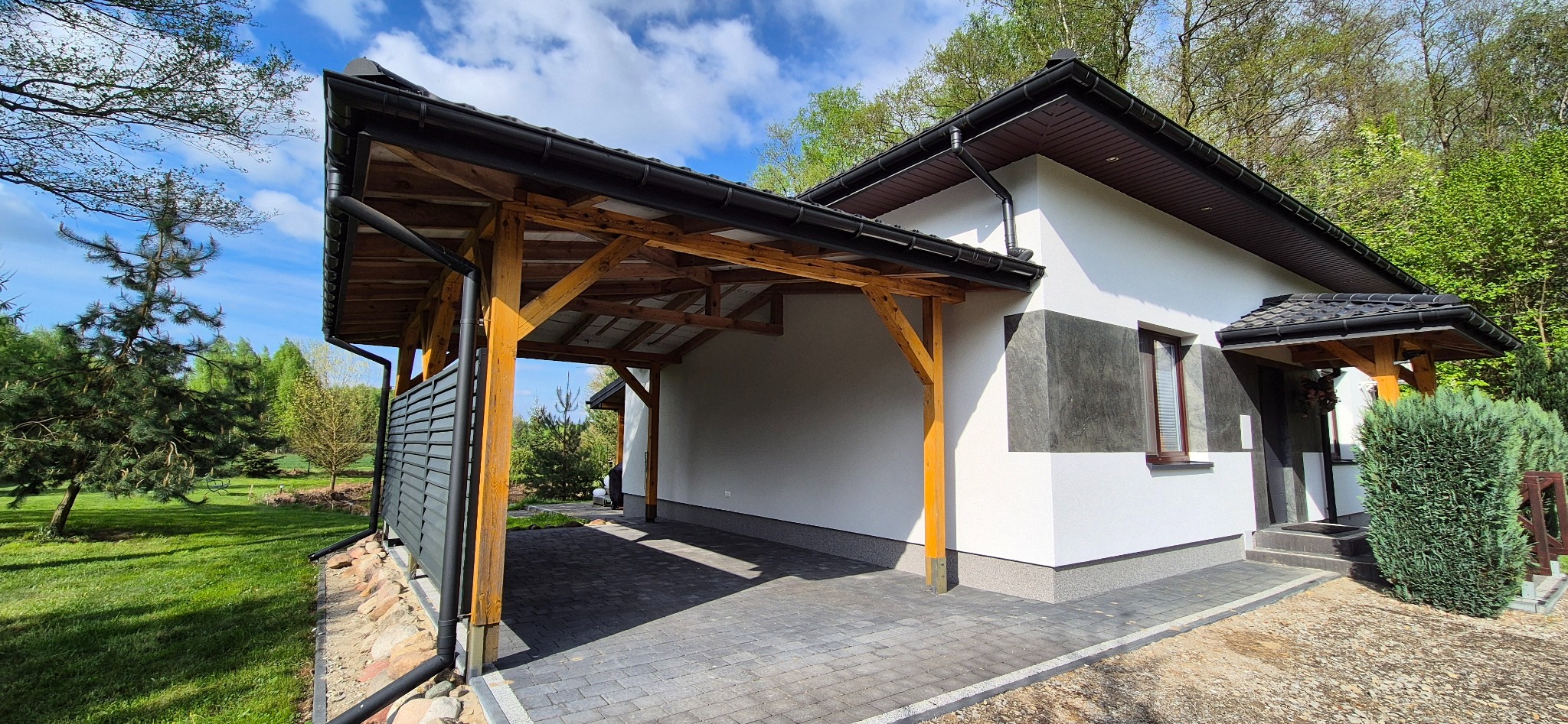 Nowoczesna elewacja domu z drewnianą konstrukcją wiaty garażowej i dekoracyjnym tynkiem w odcieniach szarości. Widok z zewnątrz, w tle zieleń drzew.