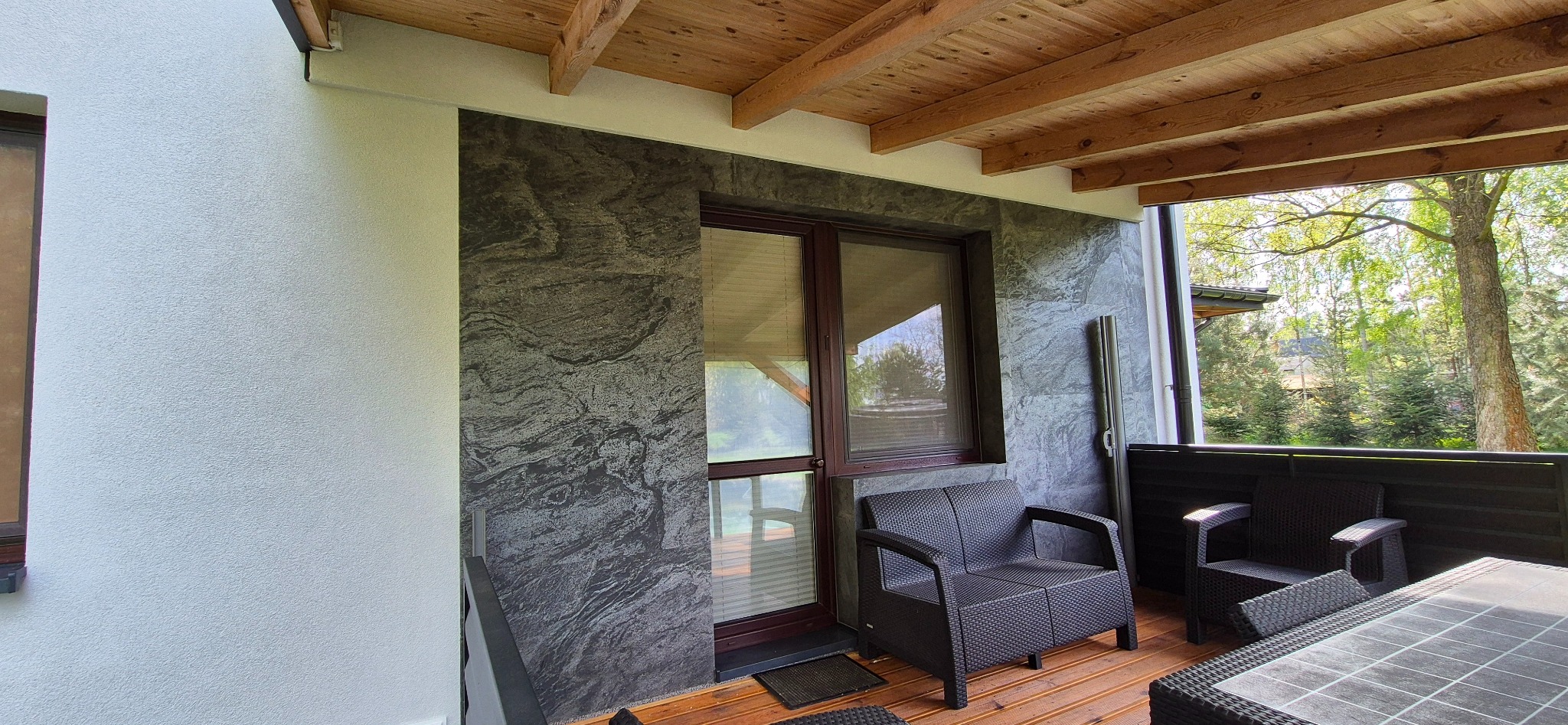 Wykończony balkon z drewnianym sufitem i ścianą z dekoracyjną szarą okładziną imitującą kamień. Widoczne meble ogrodowe i zieleń za barierką.