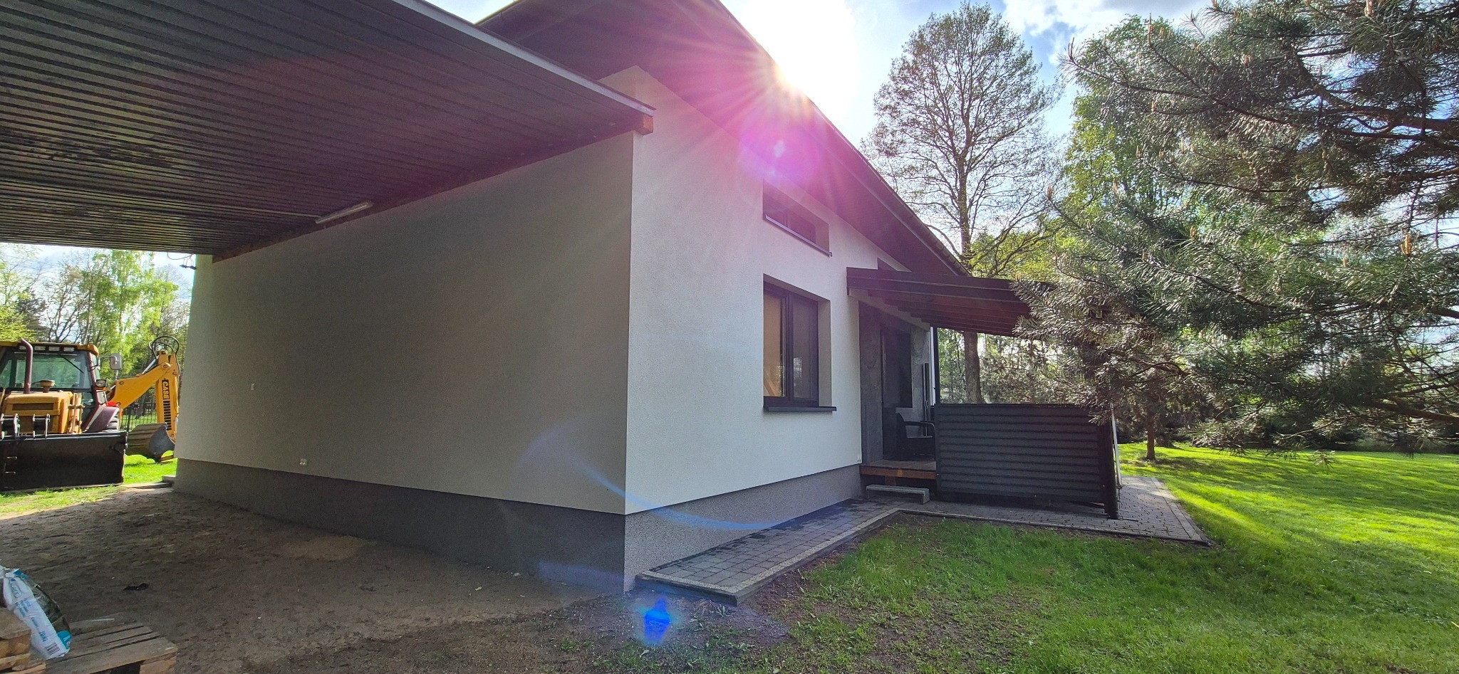 Jasny budynek z nową elewacją i podbitką dachową, widok z boku z fragmentem terenu budowy.  Na pierwszym planie trawnik. W tle drzewa i niebo.