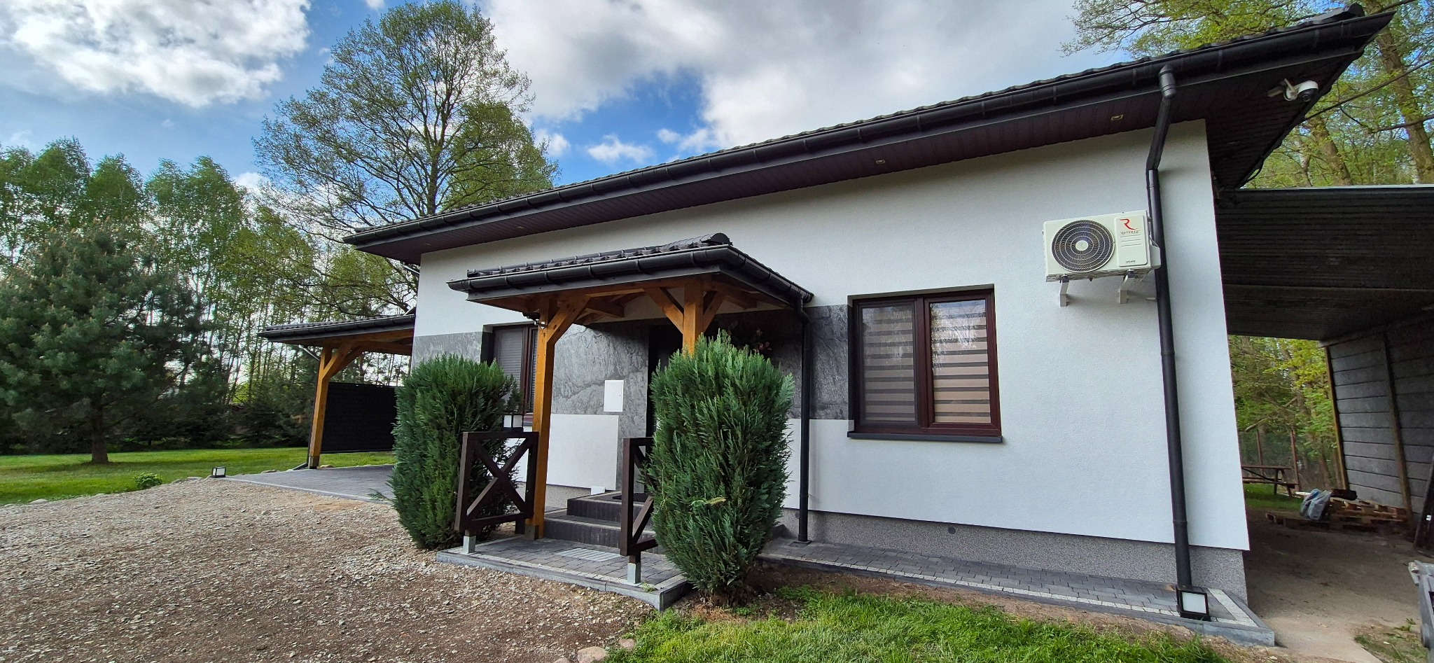 Biała elewacja domu z brązowymi oknami i drewnianym zadaszeniem nad wejściem. Widoczny klimatyzator i zadbana zieleń przed budynkiem.