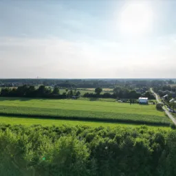 Szeroki kadr z lotu ptaka na wiejski krajobraz z polami, lasami i zabudowaniami, dominują odcienie zieleni i błękitu, jasne słońce w górnej części kadru.