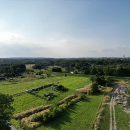 Panoramiczny widok z lotu ptaka na rozległą działkę budowlaną z częściowo wybudowanym domem i dodatkowymi budynkami gospodarczymi, otoczoną zielonymi polami i lasem w oddali, z widocznymi...