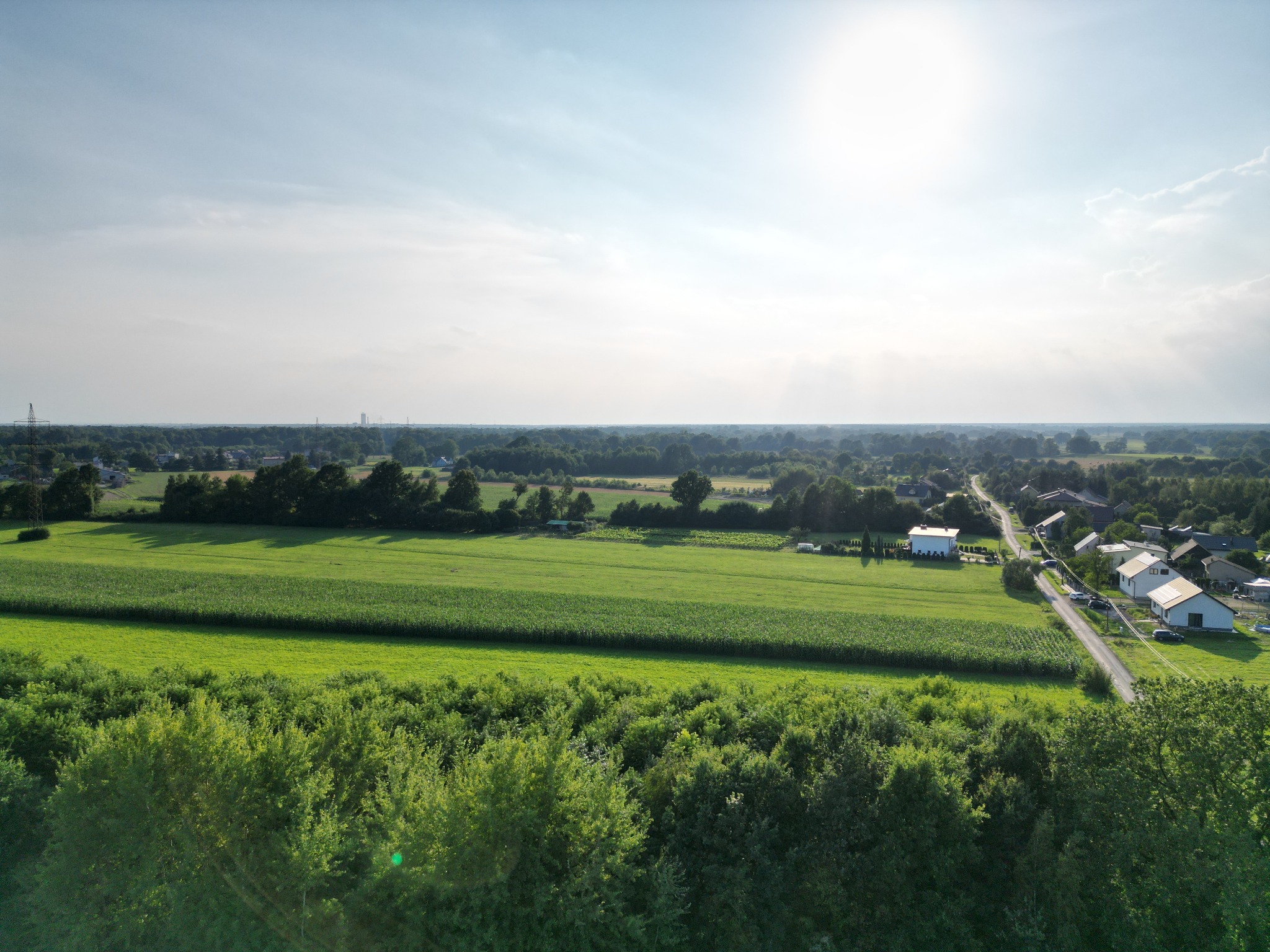 Szeroki kadr z lotu ptaka na wiejski krajobraz z polami, lasami i zabudowaniami, dominują odcienie zieleni i błękitu, jasne słońce w górnej części kadru.