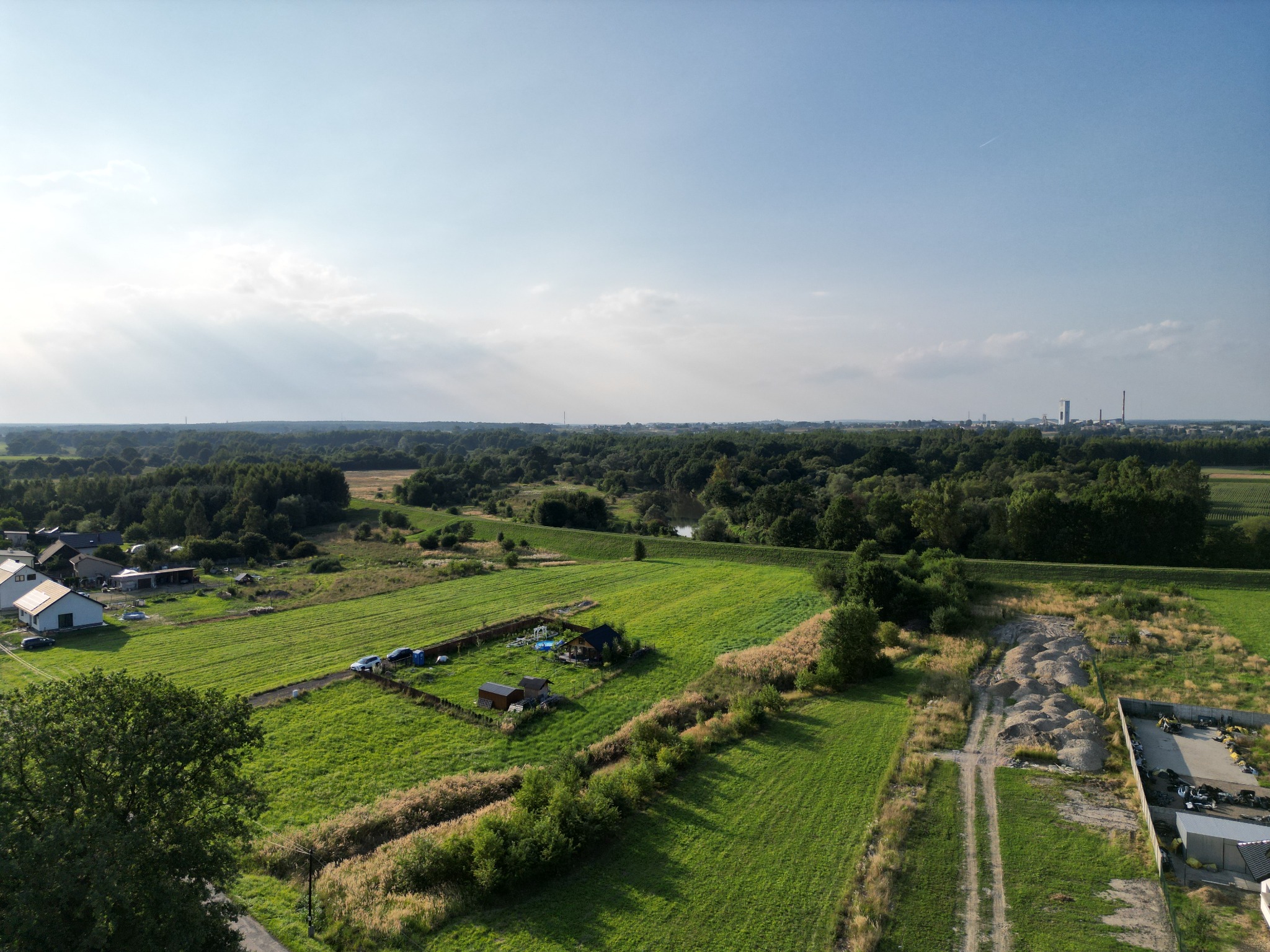 Panoramiczny widok z lotu ptaka na rozległą działkę budowlaną z częściowo wybudowanym domem i dodatkowymi budynkami gospodarczymi, otoczoną zielonymi polami i lasem w oddali, z widocznymi...