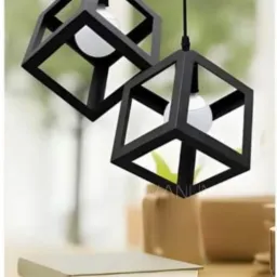 Dwa czarne, geometryczne klosze lamp wiszących w kształcie sześcianu, z żarówkami wewnątrz, zawieszone nad stołem z książkami i dekoracjami.