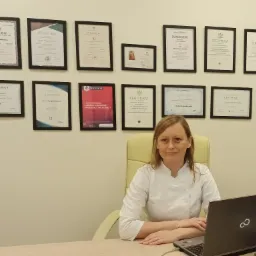 Kobieta w białym uniformie siedzi za biurkiem z laptopem, na ścianie za nią wisi kilkanaście oprawionych dyplomów i certyfikatów.