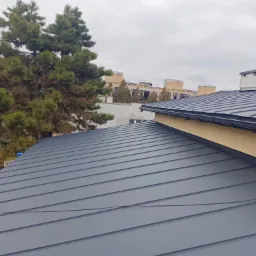 Nowy, grafitowy dach z blachy na budynku jednorodzinnym, widok z góry, na tle drzew i zabudowy miejskiej.