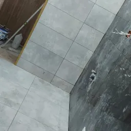 Świeżo wyłożona łazienka z szarymi płytkami imitującymi beton na podłodze i ścianach, z widocznymi puszkami elektrycznymi i wystającymi przewodami, oraz ciemnoszarą ścianą z efektem marmuru...