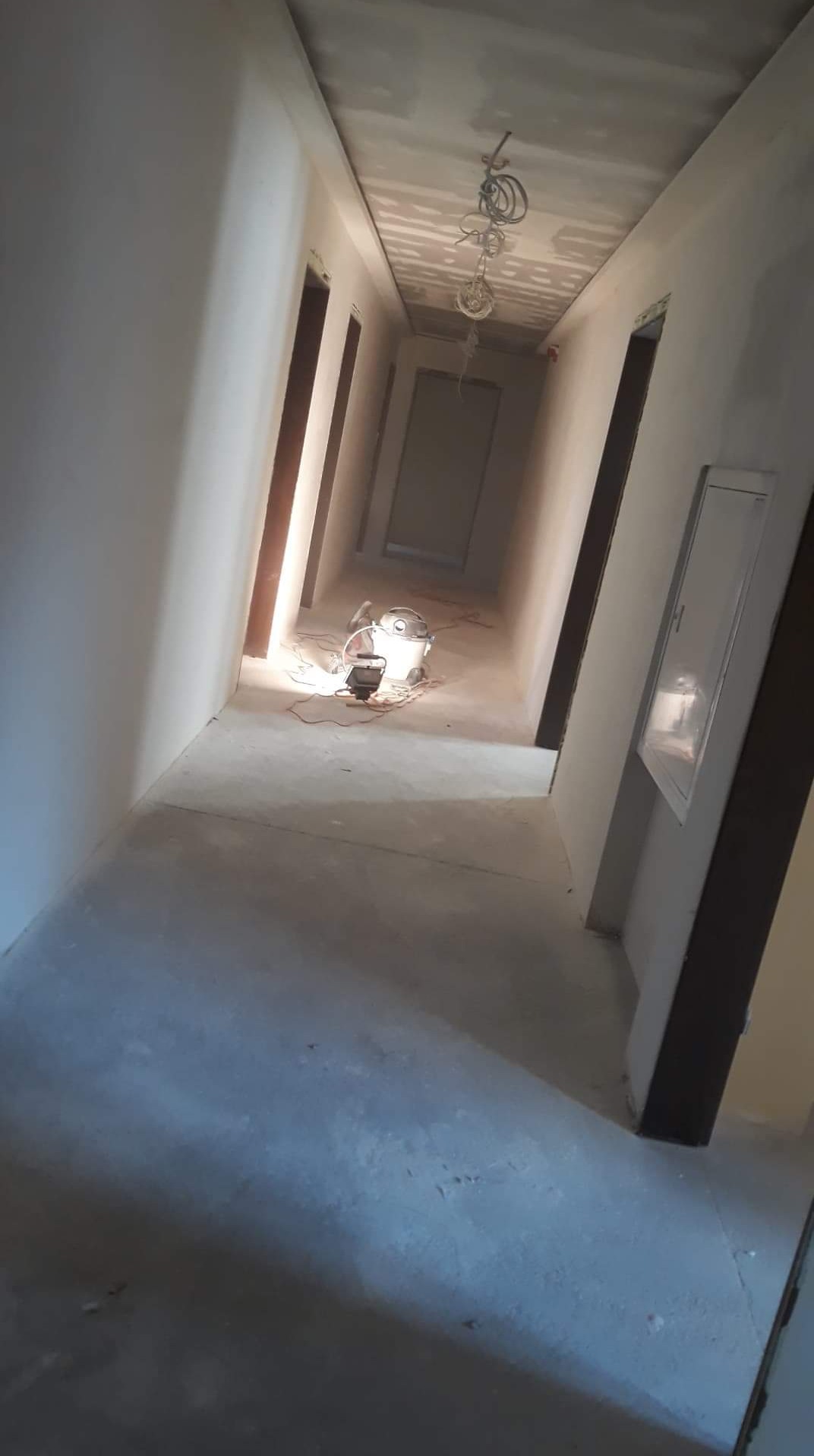 Korytarz w trakcie prac wykończeniowych, ściany przygotowane pod malowanie lub tapetowanie, widoczny sprzęt budowlany i wystające przewody elektryczne z sufitu.