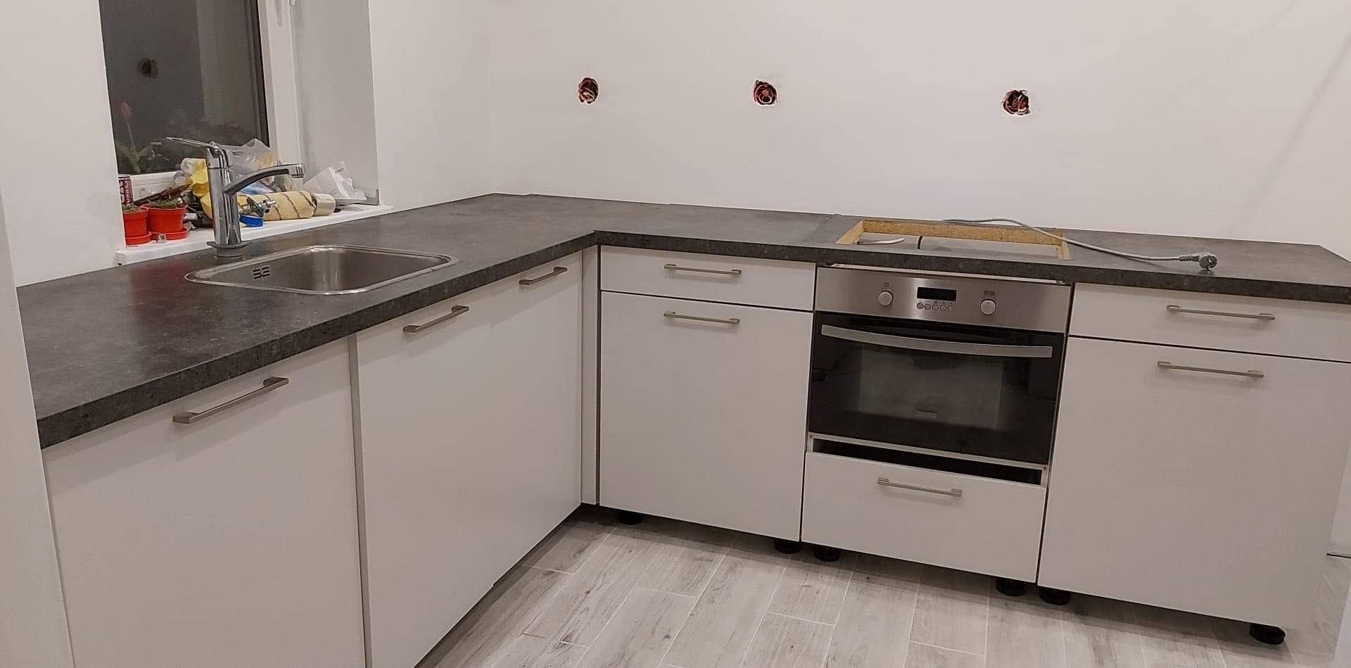 Fragment niedokończonej kuchni z białymi szafkami, szarym blatem i wbudowanym piekarnikiem. Na ścianie widoczne puszki elektryczne, a w zlewie pojemnik z płynem do naczyń.