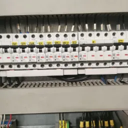 Rozdzielnica elektryczna z widocznymi bezpiecznikami i przewodami, z bliska, w szarej obudowie.