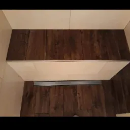 Wykończona podłoga prysznica typu walk-in z ciemnobrązowych płytek imitujących drewno i odpływem liniowym, kontrastująca z jasnymi, beżowymi płytkami ściennymi.