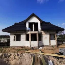 Surowy stan domu jednorodzinnego z białych bloczków, z czarnym dachem, widoczny wykop pod fundamenty, przyczepa budowlana w tle, błękitne niebo.