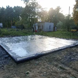 Świeżo wylana, mokra płyta fundamentowa z izolacją pionową na przygotowanym terenie pod budowę domu, widok z góry w słoneczny dzień.