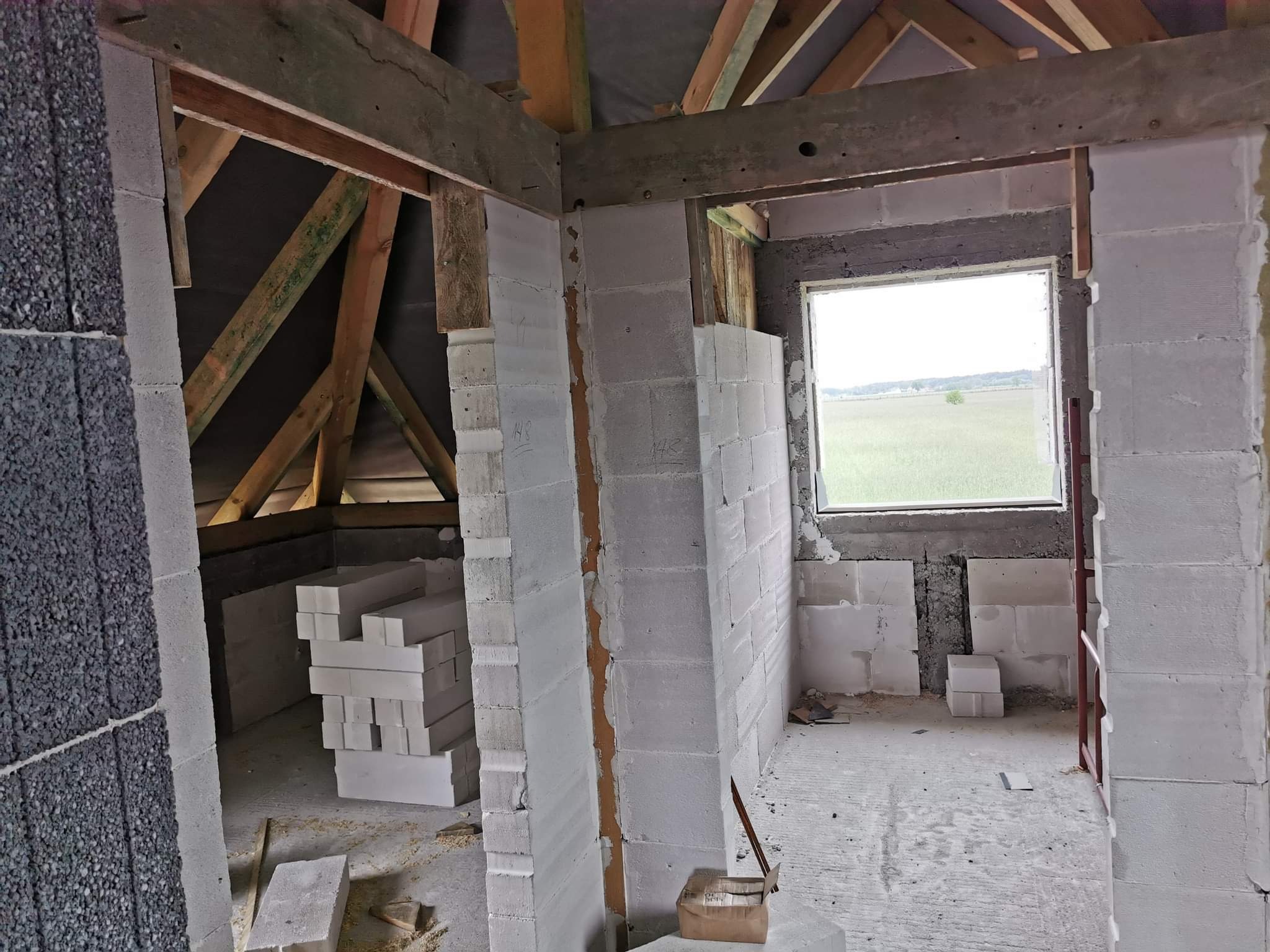 Wnętrze w surowym stanie podczas budowy domu z bloczków betonowych, widoczna konstrukcja dachu z drewnianych belek, okno z widokiem na pole, oraz stos bloczków czekających na montaż.