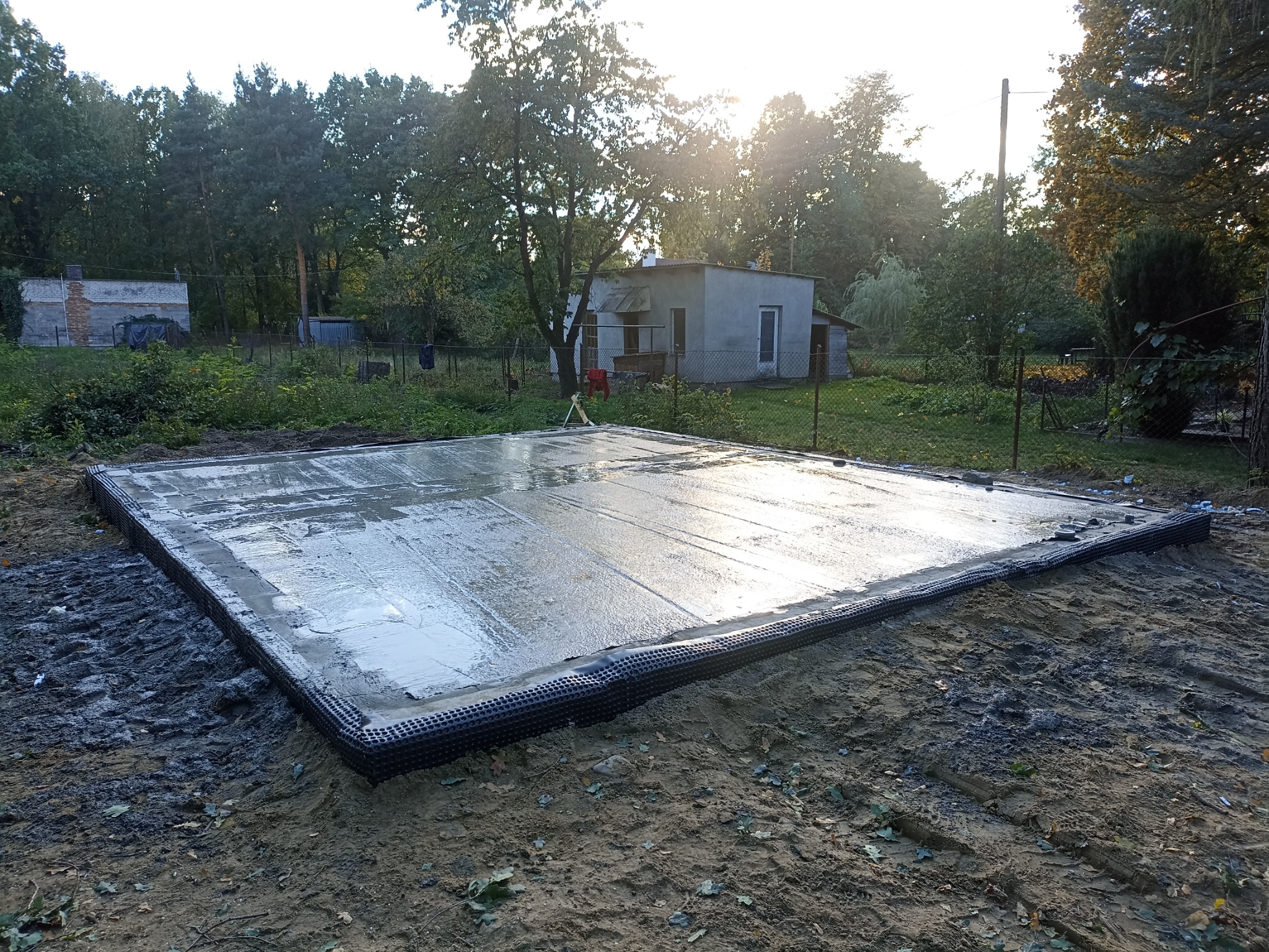 Świeżo wylana, mokra płyta fundamentowa z izolacją pionową na przygotowanym terenie pod budowę domu, widok z góry w słoneczny dzień.