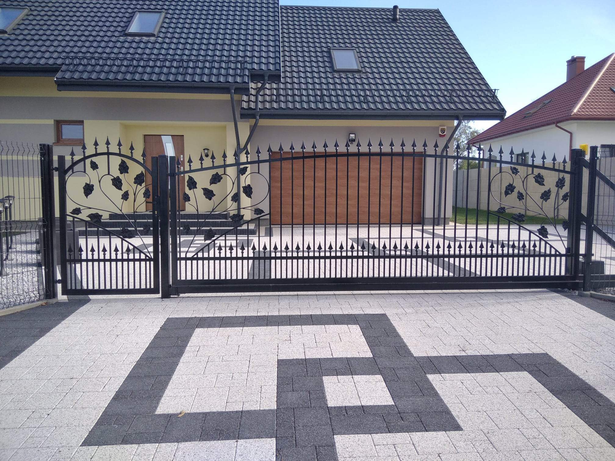 Czarna, metalowa brama wjazdowa z dekoracyjnymi elementami roślinnymi, prowadząca do nowoczesnego domu z garażem i brukowanym podjazdem we wzór geometryczny.