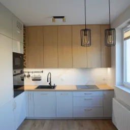 Jasna kuchnia z drewnianymi akcentami: białe szafki dolne, drewniane szafki górne, czarny zlew i bateria, drewniany panel dekoracyjny z pionowych listew, wiszące lampy w metalowych klatkach.