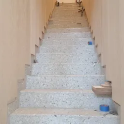 Schody wykończone szarym lastryko, z okładziną ścian z jasnego drewna, zabezpieczone niebieską taśmą malarską, w trakcie prac wykończeniowych.