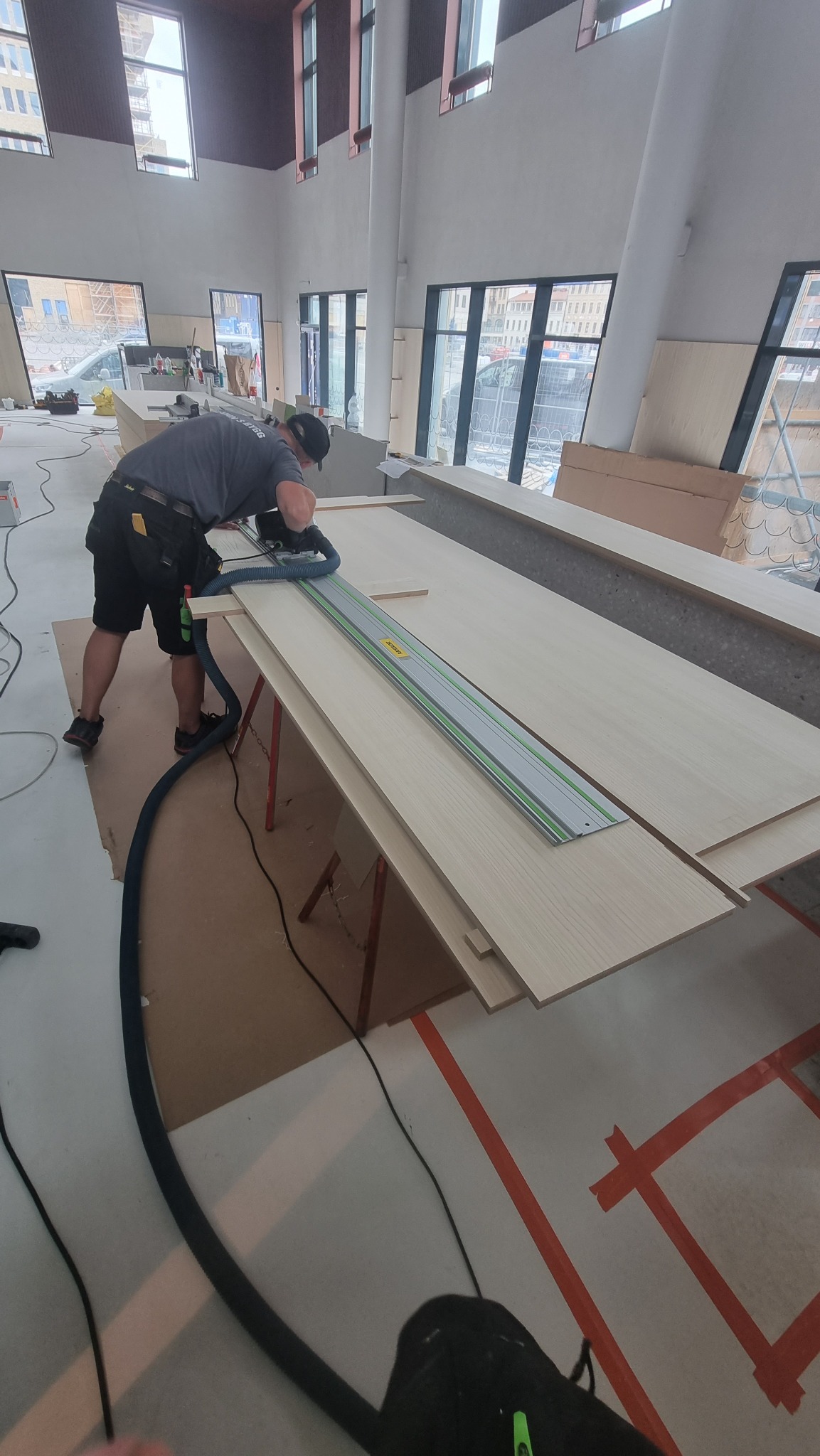 Pracownik w czarnym ubraniu tnie płytę meblową na stojaku za pomocą piły zagłębiarki z szyną prowadzącą, odkurzacz przemysłowy podłączony do piły, w tle duże okna w surowym wnętrzu.