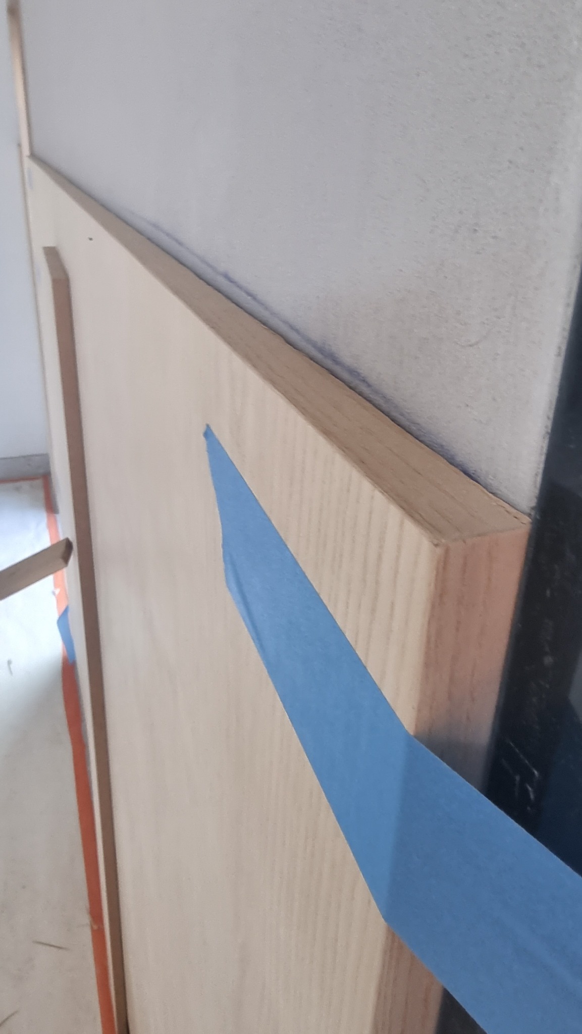 Fragment ściany z drewnianą boazerią zabezpieczoną niebieską taśmą malarską, widoczne łączenie z szarą ścianą.