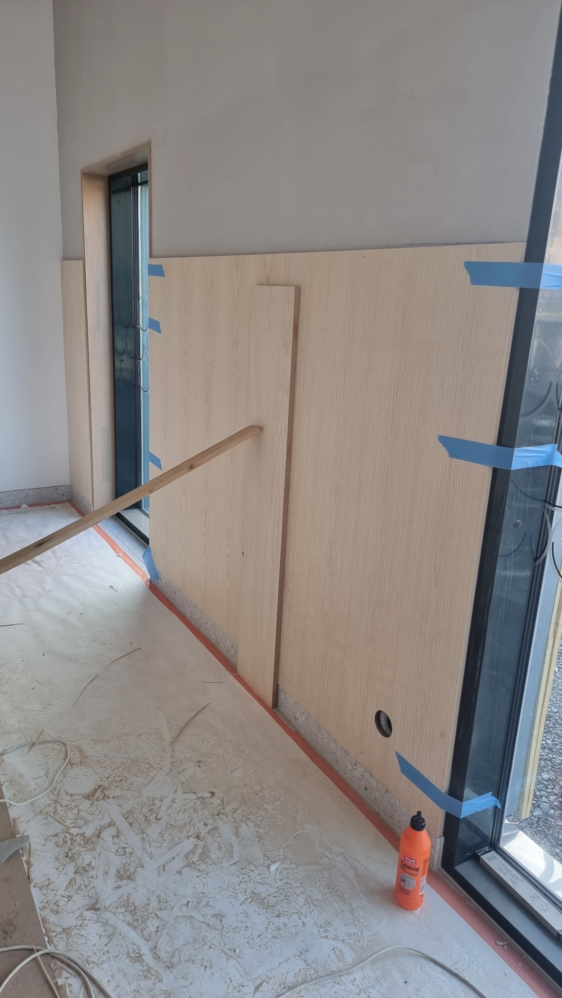 Montaż paneli ściennych z jasnego drewna, zabezpieczonych niebieską taśmą malarską, widoczna butelka kleju i fragment listwy przypodłogowej.
