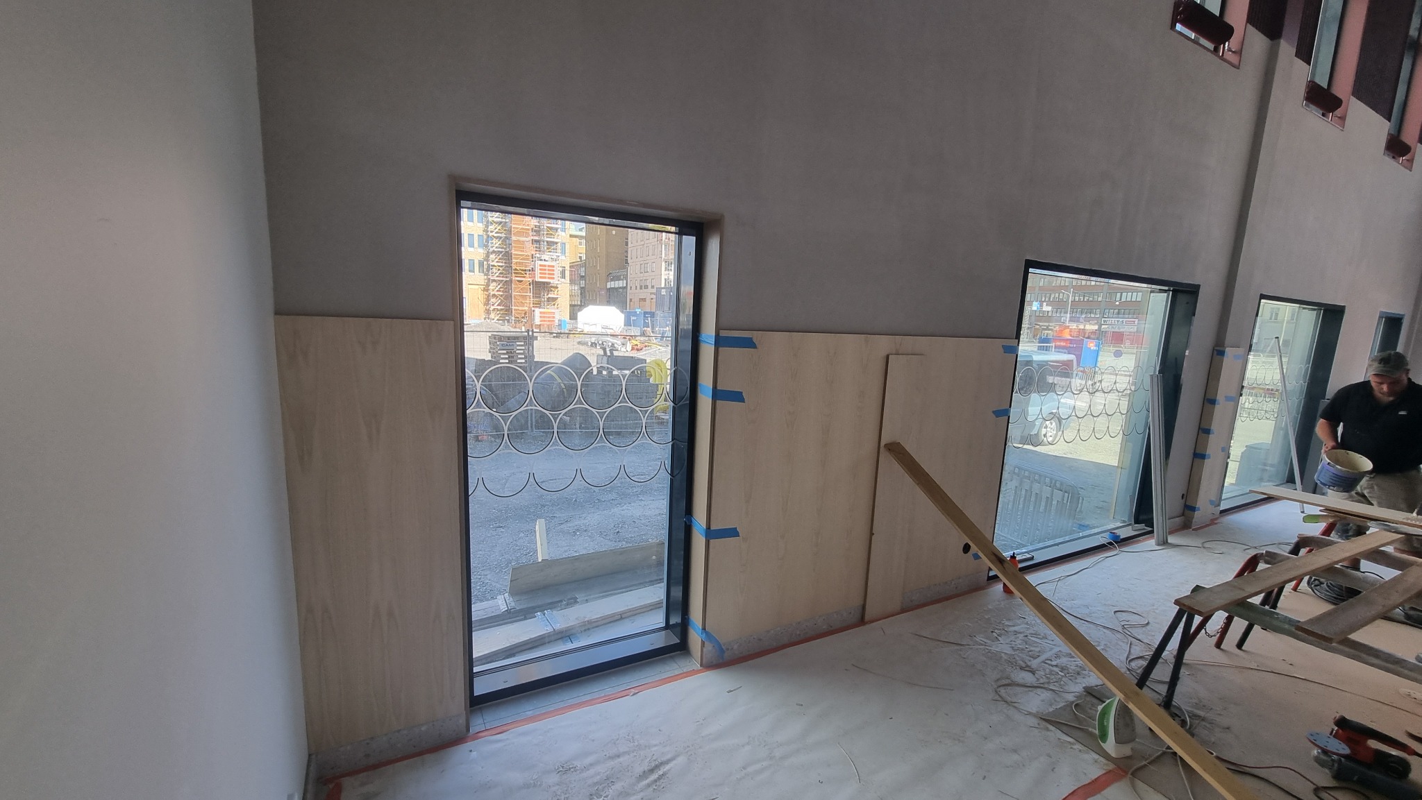 Montaż drewnianej boazerii przy oknach w surowym wnętrzu, widoczne taśmy maskujące, narzędzia i pracownik z wiadrem farby, budowa w tle za oknem.