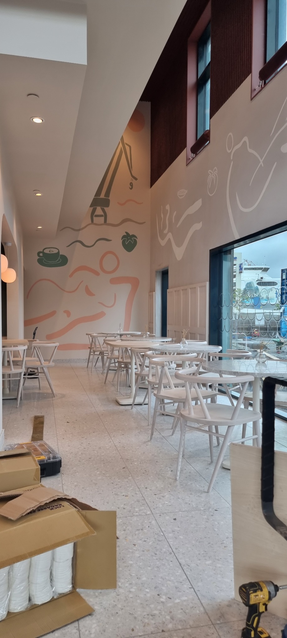 Wnętrze lokalu gastronomicznego w trakcie wykańczania: białe krzesła i stoliki z marmurowymi blatami, na ścianach abstrakcyjne malowidła, na podłodze kartony z kubkami i wkrętarka