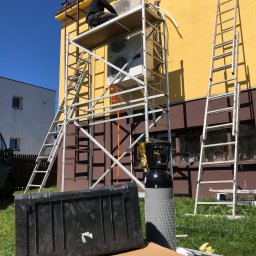 SparBud - Montaż klimatyzacji na rusztowaniu przy żółtym budynku. Widoczna butla z gazem, skrzynka narzędziowa i karton. Człowiek pracuje na górze rusztowania.