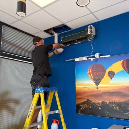SparBud - Serwisant na drabinie naprawia klimatyzator ścienny w biurze z niebieską ścianą i plakatem z balonami, widoczne narzędzia i otwarty sufit.