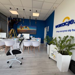 SparBud - Montaż klimatyzacji w biurze podróży: mężczyzna na drabinie instaluje urządzenie na ścianie w nowoczesnym wnętrzu z logo firmy Wakacje.pl.