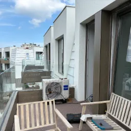 Biała jednostka zewnętrzna klimatyzacji Haier na balkonie apartamentu, narzędzia serwisowe obok, widok na nowoczesną architekturę miejską i błękitne niebo.