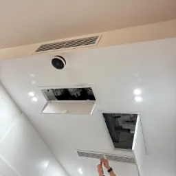 Instalator w trakcie montażu klimatyzacji kanałowej w suficie podwieszanym, widoczne kratki wentylacyjne i otwory rewizyjne. Białe wnętrze, oświetlenie punktowe.