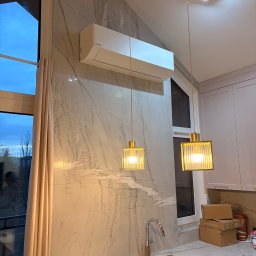 SparBud - Biała klimatyzacja ścienna w nowoczesnej kuchni z marmurową ścianą, złotymi lampami i oknem z widokiem na zewnątrz. Minimalistyczny design.