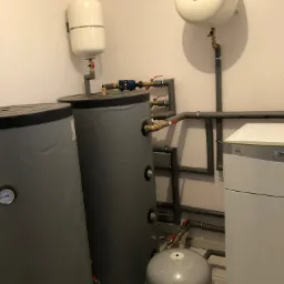 Instalacja z dwoma szarymi zbiornikami akumulacyjnymi ciepła, białym zasobnikiem ciśnieniowym, rurami w izolacji i kotłem grzewczym Vaillant Green iQ na białej ścianie, z widocznymi zaworami...