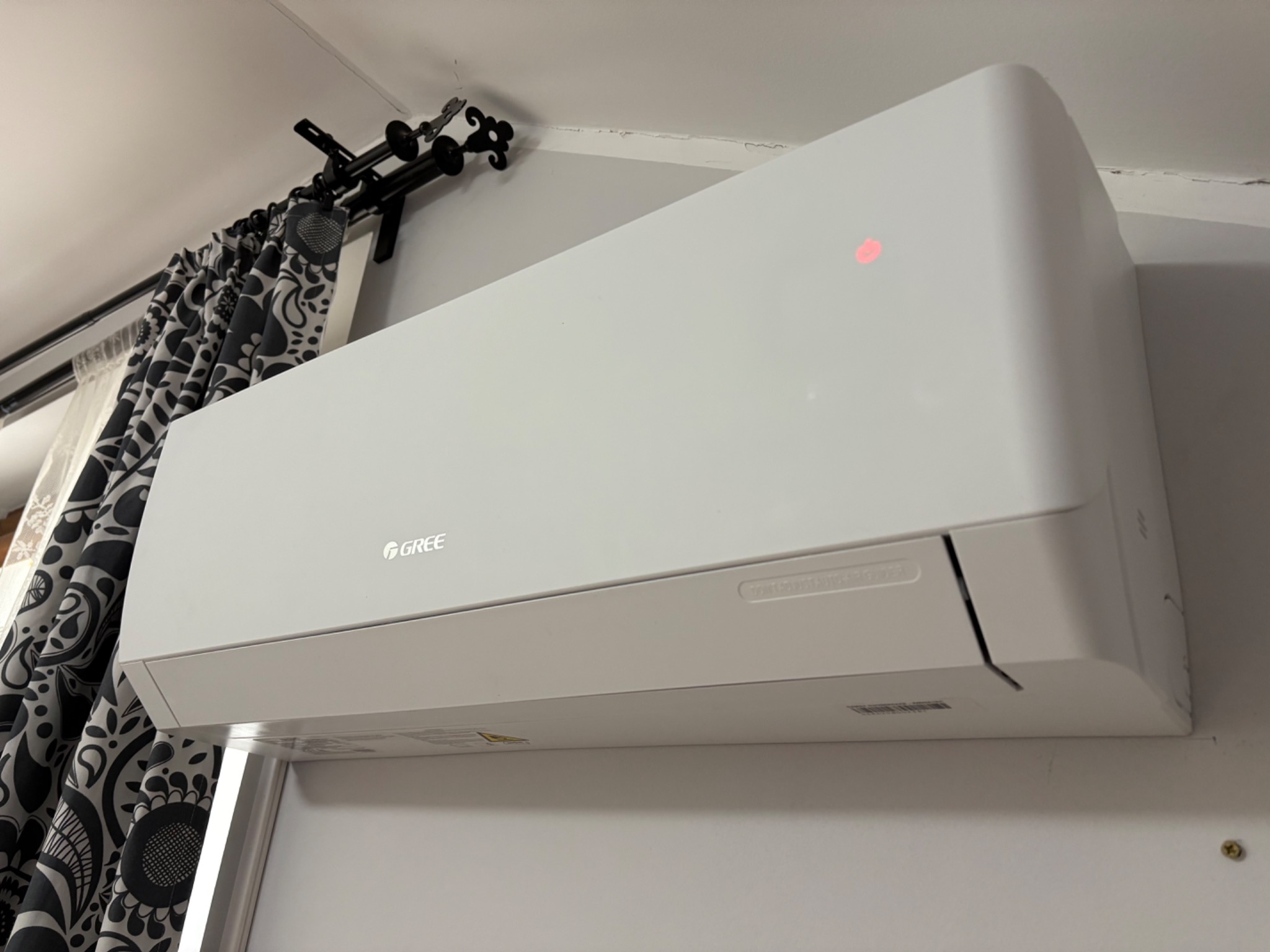Biały klimatyzator marki Gree zawieszony na ścianie, widoczny fragment zasłony z wzorem i karnisz.